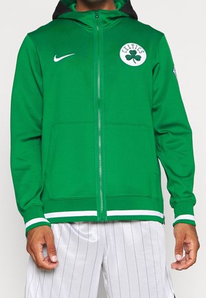 Homme portant un sweat à capuche zippé vert avec le logo des Celtics et le swoosh blanc de Nike, associé à un short blanc à fines rayures.