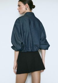 Dunkelbluse aus Denim mit gerafftem Saum und voluminösen Ärmeln, kombiniert mit einem schwarzen Minirock. Glatte Textur mit einem entspannten Design.