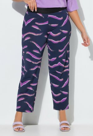 Donna che indossa pantaloni blu navy con motivi astratti a pennellate viola e rosa, un top lavanda e sandali lavanda aperti in punta.
