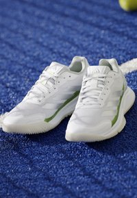 Scarpe sportive bianche con dettagli verdi su una superficie sportiva blu testurizzata vicino a una linea bianca e una palla parzialmente gialla.