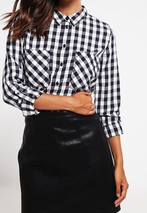 Kobieta ubrana w czarno-białą koszulę w kratę z podwiniętymi rękawami i czarną skórzaną spódnicę, trzymająca się za talię jedną ręką.