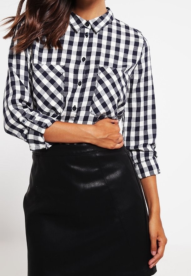 Femme portant une chemise à carreaux noire et blanche boutonnée avec les manches retroussées et une jupe en cuir noire, tenant sa taille d'une main.