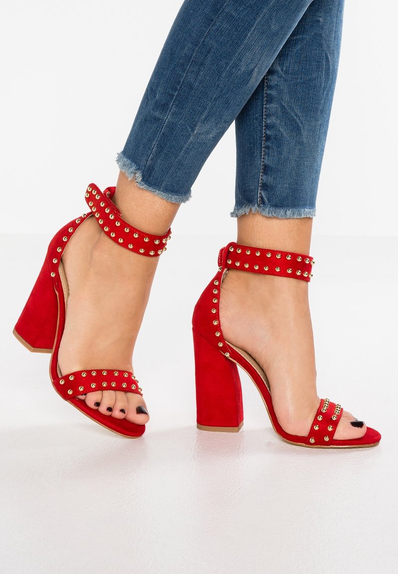 Sandalias de tacón alto de ante rojo con correas en el tobillo, adornadas con tachuelas doradas, con punta abierta y un tacón grueso. Combinadas con jeans de dobladillo deshilachado.