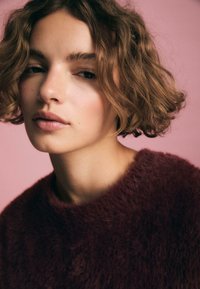 Weinrote, flauschige Sweater mit rundem Ausschnitt. Kurze, wellige braune Haare rahmen das Gesicht ein. Der sanfte rosa Hintergrund hebt die weichen Züge hervor.