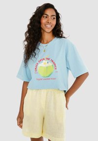 Modré tričko s krátkým rukávem s grafikou kokosu a textem. V páru se světle žlutými šortkami, oboje vyrobené z měkké látky.
