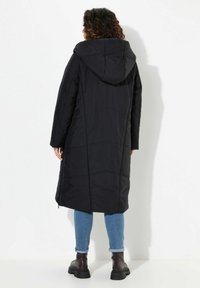 Svart vadderad jacka med huva, med en lång och rak design och quiltad textur. Bärs över blå jeans och svarta klumpiga stövlar.