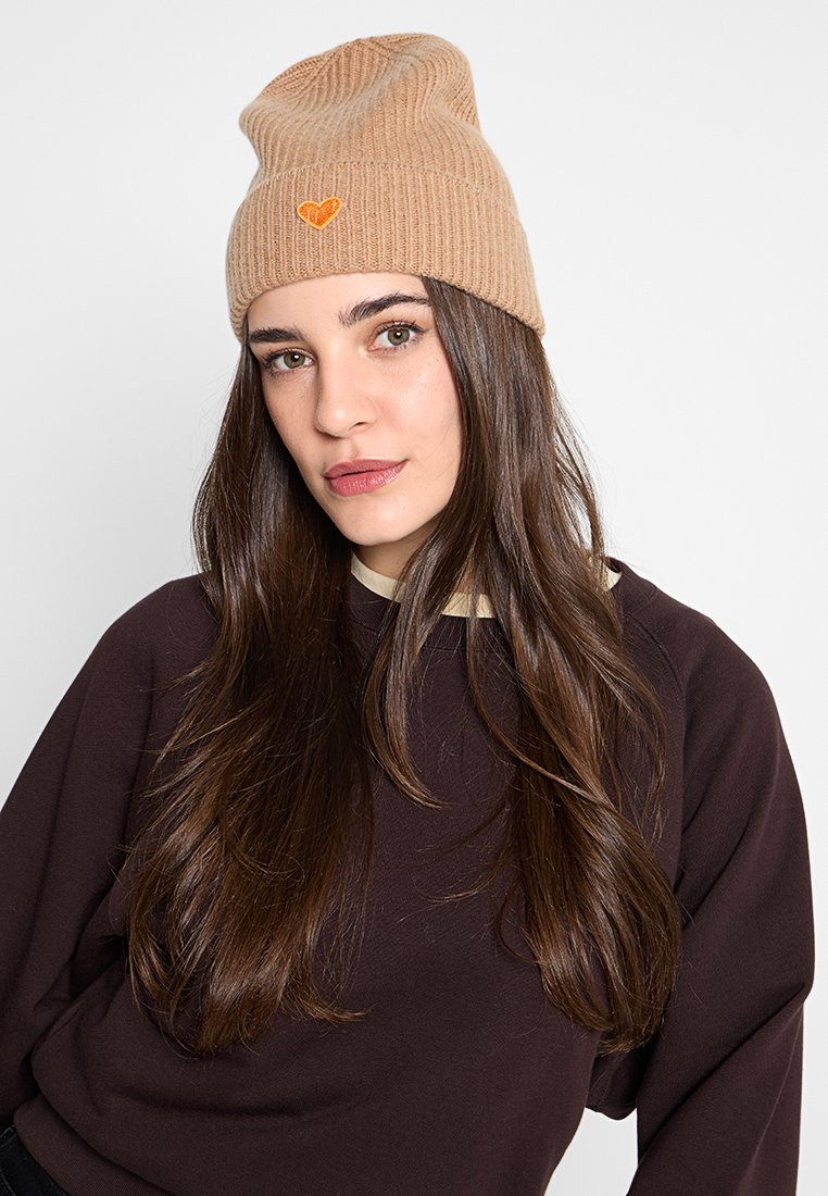 Młoda kobieta z długimi ciemnymi włosami, nosząca beżową dzianą czapkę z pomarańczowym sercem oraz ciemnobrązowy sweter na tle białym.