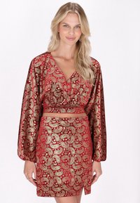 Ein rotes und goldenes Paisleymuster-Outfit mit einem bauchfreien Oberteil, das lange Ärmel hat, und einem passenden Rock mit einem seitlichen Schlitz.