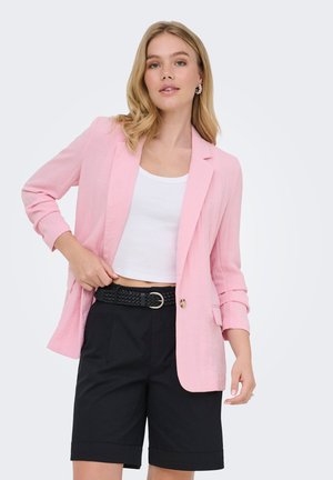 Woman wearing a light pink blazer over a white crop top, black belted shorts, and decorative earrings, posing with one hand in pocket.