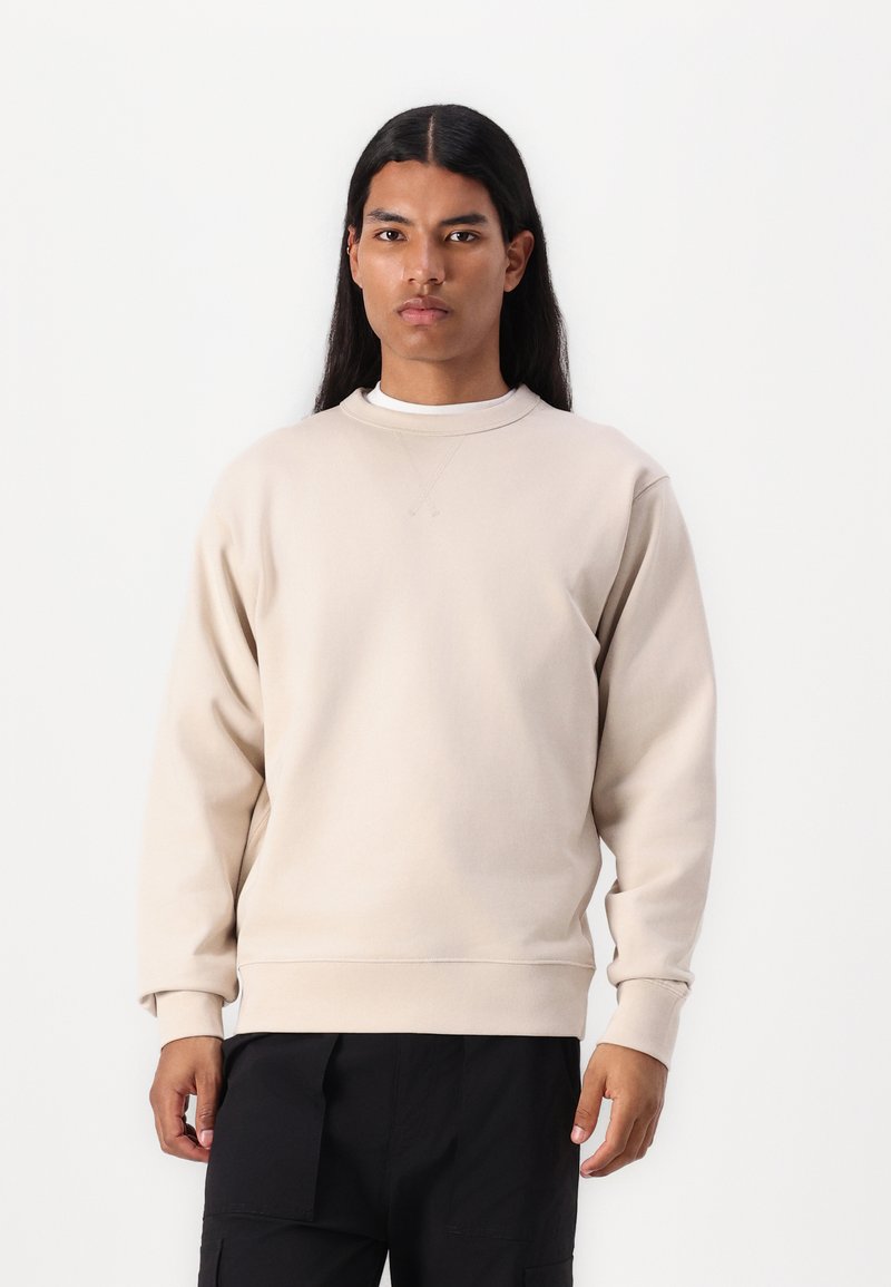 Sweatshirt bege com decote redondo e mangas compridas, feito de tecido macio. Apresenta punhos e bainha canelados, com detalhes de costura discretos.