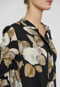Melna blūze ar ziedu rakstu, kurā ir krēmkrāsas un brūnas puķes. Dizainā ir pogu ailes un apkakle, izgatavota no viegla auduma.