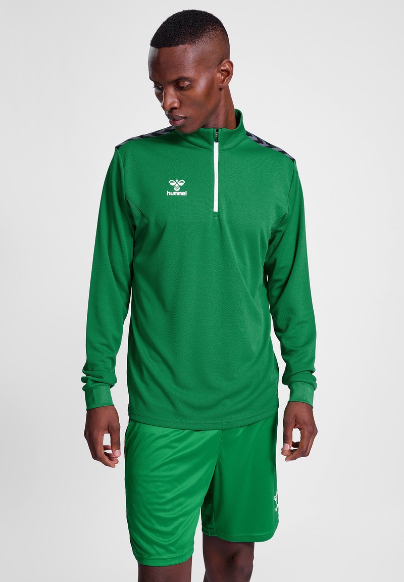 Grøn atletisk langærmet top med halvmund-zipper og sorte mønstrede accenter, parret med matchende grønne shorts. Lavet af letvægts, åndbart stof.