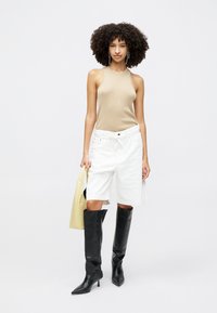 Bodysuit neule beige, shorts denim valkoiset ja mustat polven ylle ulottuvat kärjet saappaat. Malli pitää vaaleankeltaista olkalaukkua. Yksinkertainen valkoinen tausta.