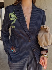 Tamsiai mėlynas dryžuotas švarkas, papuoštas žaliu gyvatės siuvinėjimu ant apykaklės ir saulės grafika kairėje kišenėje. Rankoje laikoma dramblio kaulo spalvos delninė.