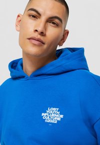 Blå hettegenser i bomullsblanding, med hvit trykt logo-tekst "LOST YOUTH INFLUENCE CULTURE ©2022" på brystet.