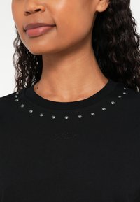 Giovane che indossa una maglia nera con borchie metalliche intorno allo scollo e un testo sottile ricamato sul petto.