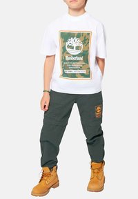 T-shirt blanc avec un graphisme Timberland vert et orange, associé à un pantalon cargo vert foncé avec des poches et un logo orange, et des bottes de travail beiges.
