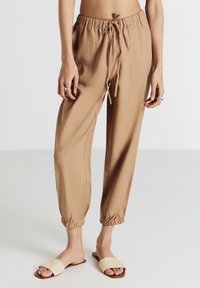 Persona che indossa pantaloni jogger beige con coulisse e polsini elasticizzati, in piedi su un pavimento chiaro contro uno sfondo bianco.