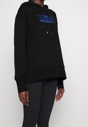 Osoba ubrana w czarną, oversize'ową bluzę z kapturem z niebieskim logo na piersi oraz czarne dopasowane legginsy na jednolitym tle.