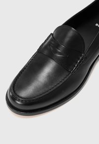 Zwarte leren loafers met een gladde afwerking, voorzien van stiksels rondom de teen en een decoratieve band aan de voorkant.