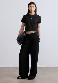 Μαύρο cropped t-shirt με μικρό λευκό λογότυπο, συνδυασμένο με μαύρο παντελόνι ψηλόμεσο και φαρδύ σε ίσια γραμμή. Το μοντέλο κρατά μια μαύρη δερμάτινη τσάντα.