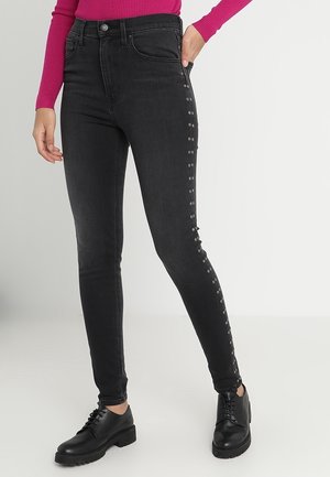 Mujer vistiendo jeans skinny de cintura alta negros con detalles de ojales metálicos en los laterales, zapatos negros con cordones y una blusa de manga larga rosa brillante.