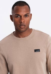Felpa beige con scollo rotondo, tessuto testurizzato e un patch logo rettangolare nero sul petto.
