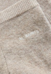 Tejido de punto beige con una textura suave, que presenta un sutil logo de AllSaints bordado en blanco. Vista en primer plano, mostrando el detalle del tejido.