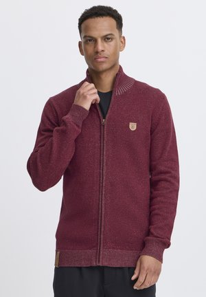Sudadera burdeos con cremallera y cuello alto, puños acanalados y un pequeño parche logotipo en el pecho. Tejido de punto texturizado con un ligero brillo.