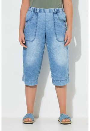 Person iført lyseblå denim knælange shorts med store forlomme, en grøn top og blå sandaler med åben tå, står op ad en væg.