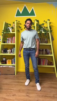 Jasnozielona koszulka z krótkim rękawem, niebieskie dżinsy i białe trampki. W tle żółte ściany, zielone półki i rośliny doniczkowe.