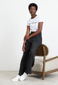 Witte t-shirt met "Calvin Klein Jeans" print, gecombineerd met losse zwarte broek en witte sneakers, zittend op een lichthouten stoel.