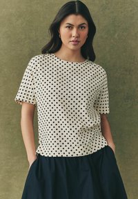 Kortærmet beige t-shirt med sorte prikker, rund halsudskæring, afslappet pasform, struktureret stof. Kombineret med en marineblå nederdel, hænderne i lommerne.