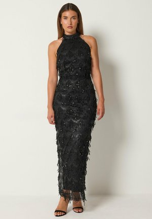 Donna che indossa un abito nero senza maniche con paillettes, dettagli di frange e collo alto, in piedi davanti a uno sfondo chiaro e uniforme.