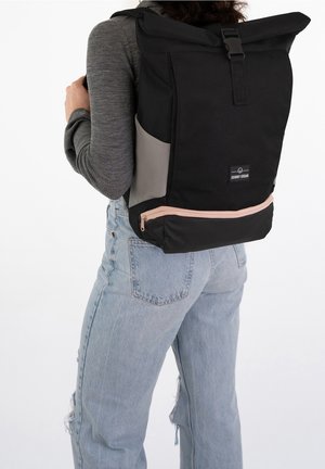 Persona che indossa uno zaino a rotolo nero e grigio con tasca con cerniera rosa, maglia grigia a maniche lunghe e jeans blu chiaro strappati, di spalle.