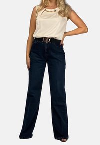 Kremowy bezrękawnik z łańcuchowym akcentem, zestawiony z ciemnoniebieskimi jeansami o wysokim stanie i szerokich nogawkach oraz czarnym paskiem ze złotą klamrą.