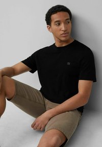 Schwarzes T-Shirt mit kurzen Ärmeln und Rundhalsausschnitt sowie einem kleinen Logodetail. Getragen mit khakifarbenen Shorts. Glatter Stoff, lässige Passform.