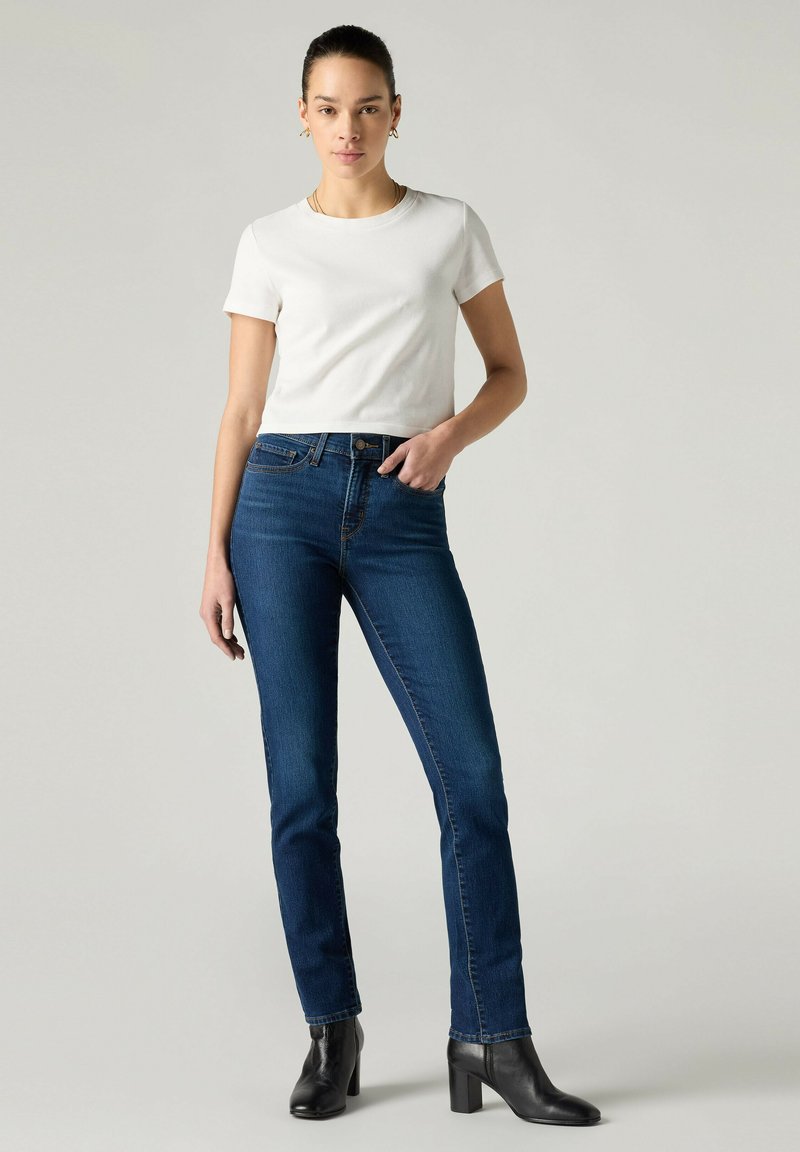 Femme debout portant un t-shirt blanc à manches courtes, un jean skinny taille haute bleu foncé et des bottines noires à talons sur un fond uni.