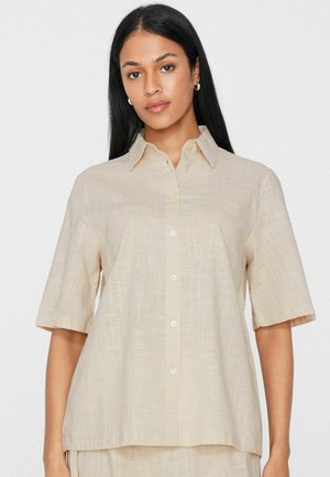 Frau mit langen schwarzen Haaren, die ein beiges kurzärmeliges Hemd mit Kragen vor einem schlichten Hintergrund trägt.