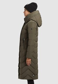 Vrouw in een lange olijfgroene gewatteerde jas met capuchon en zwarte beanie, staand in profiel tegen een neutrale achtergrond.