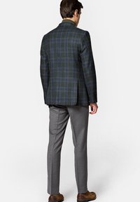 Zielona marynarka w kratę z wyciętym kołnierzem, dopasowany krój i szare spodnie. Marynarka ma niebieskie i brązowe akcenty oraz teksturowaną tkaninę.