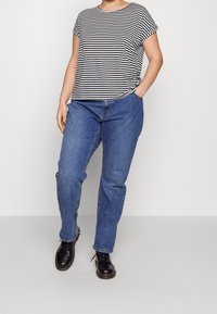 Camiseta de rayas en blanco y negro de manga corta combinada con unos jeans de mezclilla azul claro y zapatos negros de cordones, contra un fondo sencillo.