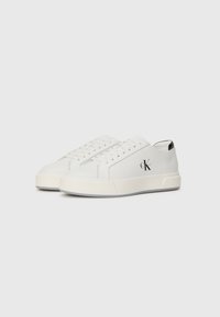Adidași albi din piele netedă, cu vârf rotund, design cu șireturi, talpă groasă din cauciuc, accentuați cu un logo negru pe lateral, stil minimalist.