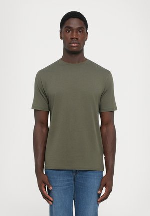 Mann mit kurzen Haaren, der ein schlichtes olivgrünes T-Shirt mit kurzen Ärmeln und blaue Jeans trägt, steht vor einem weißen Hintergrund.
