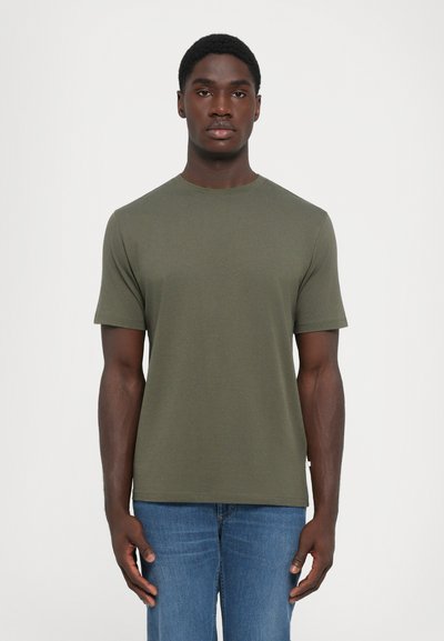 Homme à cheveux courts portant un t-shirt uni vert olive à manches courtes et un jean bleu, debout devant un fond blanc.