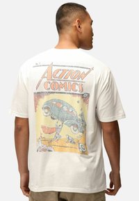 Weißes Baumwoll-T-Shirt mit einer retro "Action Comics" Grafik, die farbenfrohe Illustrationen von Superhelden und einem Auto zeigt.
