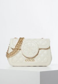 Tikkipintainen kermanvärinen käsilaukku kultainen ketjuhihna, jossa pyöreä merkki ja kohokuvioitu logo edessä. Sileä pinta ja jämäkkä muoto.