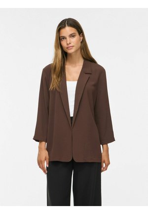 Kvinde med langt brunt hår iført en løs brun blazer over en hvid top og sorte bukser, stående mod en ensfarvet lys baggrund.