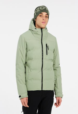 Chaqueta acolchada verde claro con cremallera frontal, bolsillos laterales y capucha ajustable. Combinada con un gorro verde oscuro que tiene un logotipo.
