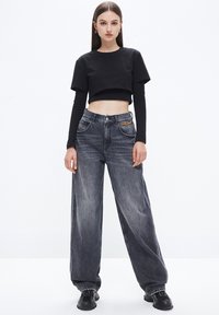 Camiseta negra corta en capas sobre una blusa negra de manga larga, combinada con jeans oscuros de corte holgado y detalle de cadena dorada.